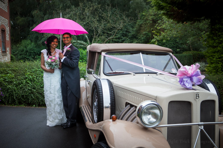bride and groom under an umbrella with wedding car